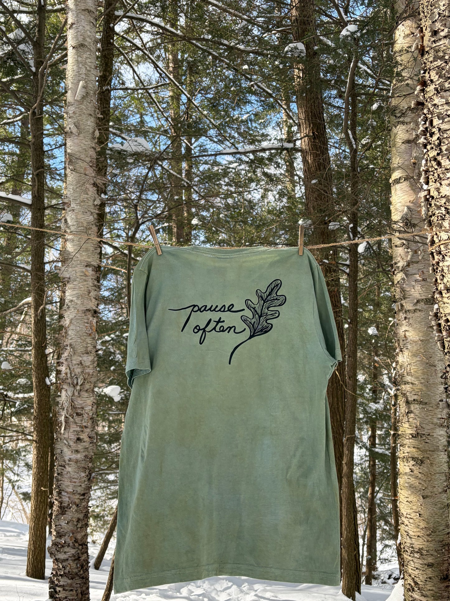 Green shirt with 'Pause Often' text hanging between trees in forest. Ecology t-shirt.