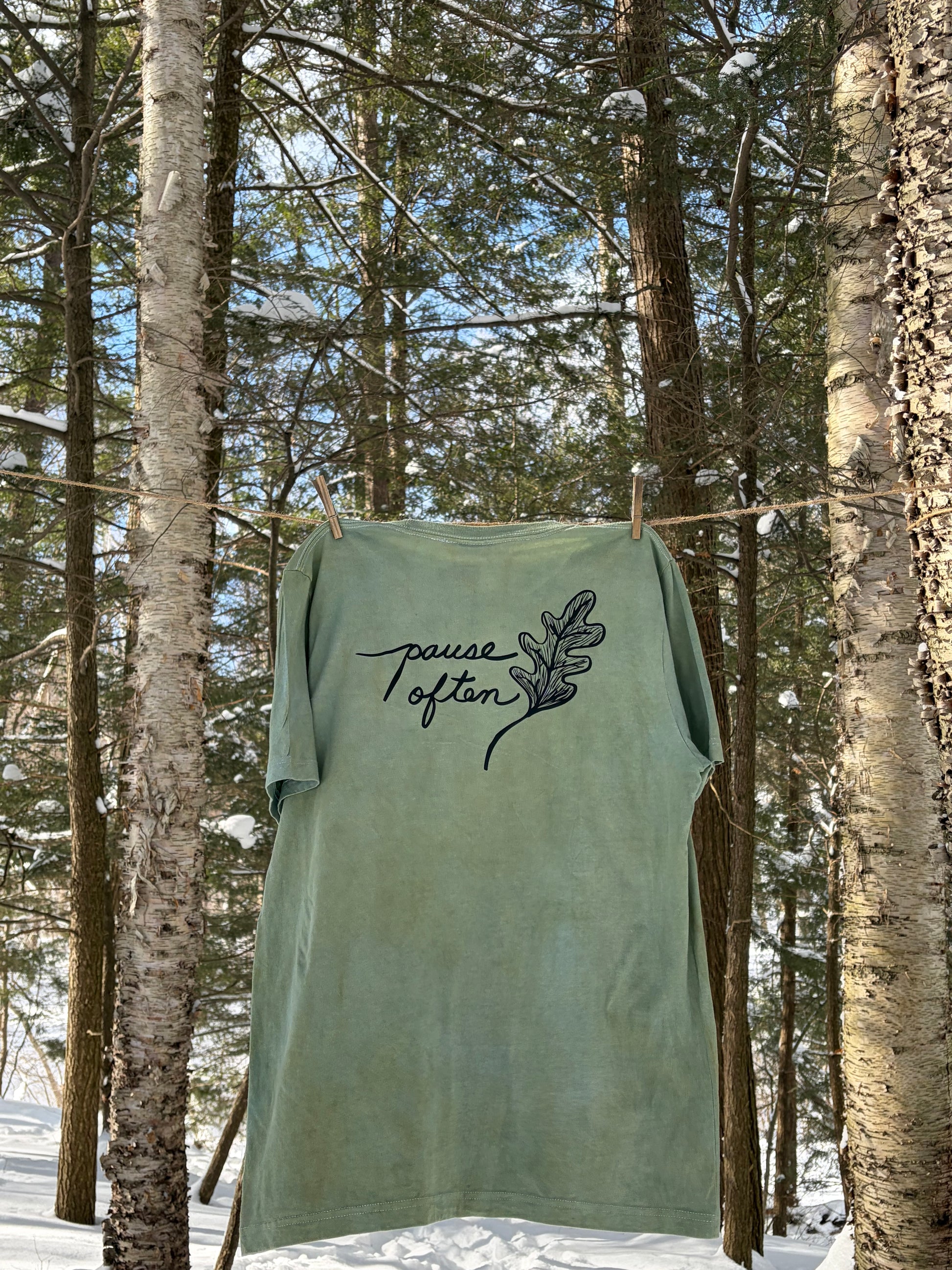 Green shirt with 'Pause Often' text hanging between trees in forest. Ecology t-shirt.