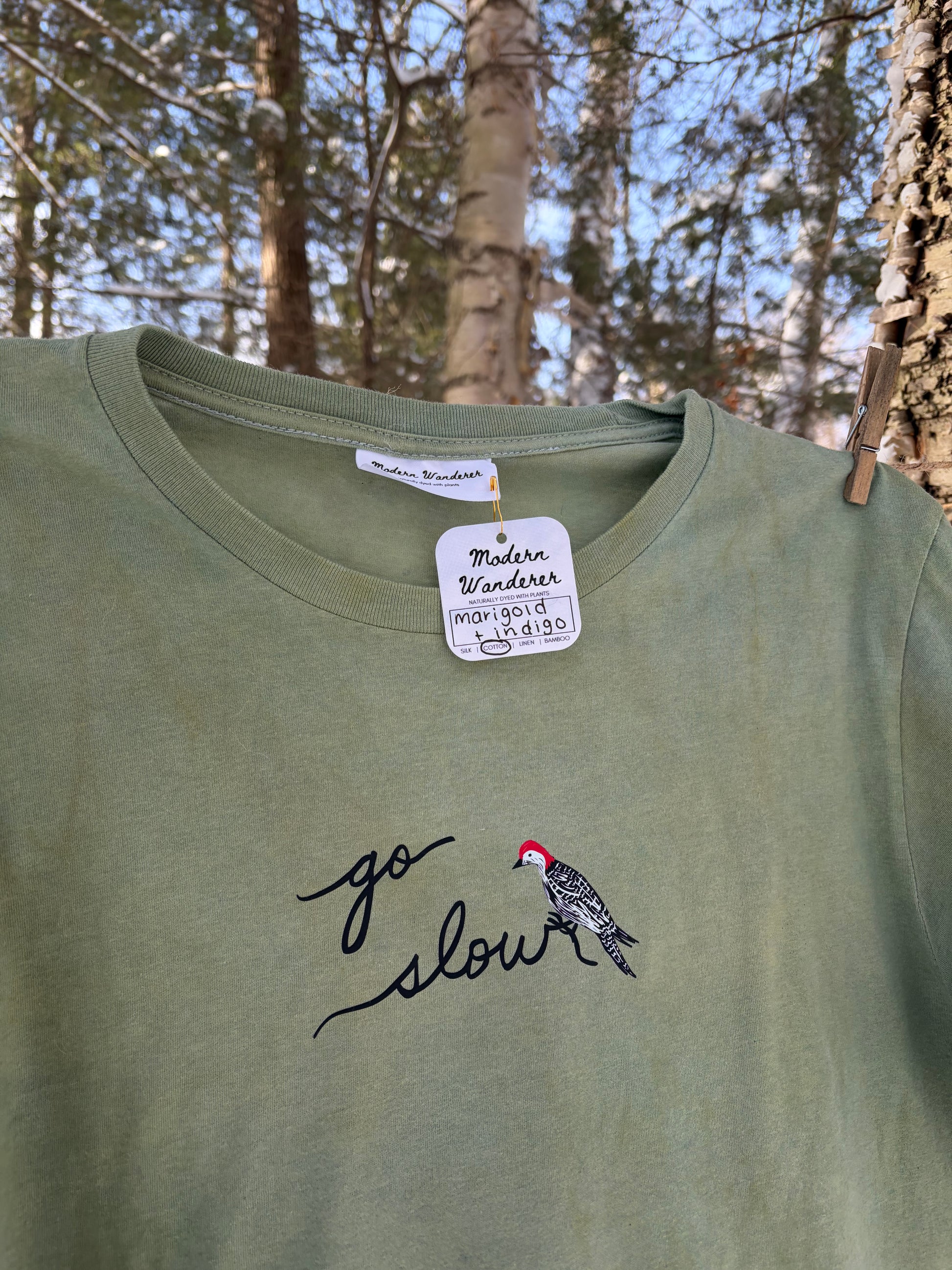 Green t-shirt with a bird design and 'go slow' text, hanging outdoors with trees in the background. Plant dyed.